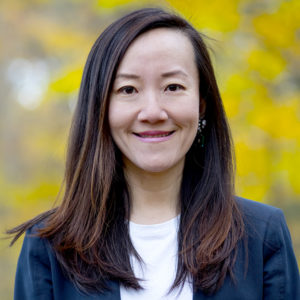 Head and shoulders images of a brunette woman smiles at camera with blue blazer and white t-shire outside with a blurred yellow background.