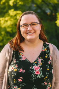 Caucasian woman with brown hair and glasses smiles at camera. She's wearing a tan cardigan and black tank top with pink and yellow flowes.