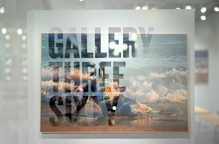 Photo peers through the glass of the Gallery 360 walls, looking at Joiri Minaya's "Container #6" piece, which shows a woman reclining in a full body suit on the beach.