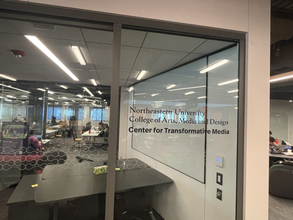 A glass-walled classroom or study space at Northeastern University. The wall features text reading 'Northeastern University College of Arts, Media and Design Center for Transformative Media'. Inside the room, students can be seen working at tables under fluorescent lighting. The space has a modern, institutional design with dark flooring and white walls.