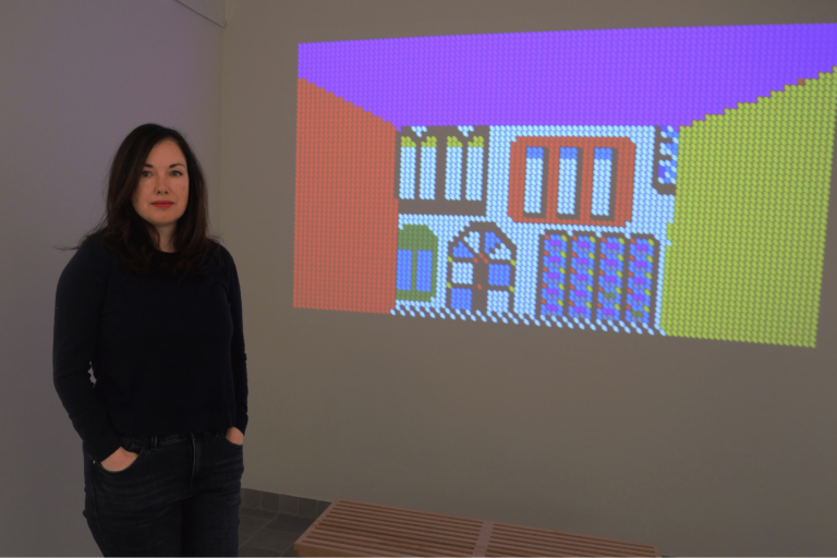 Portrait of Cat Mazza by Jim Finn. She has a serious expression and is wearing a black shirt with her hands in her pockets. In the background, her video piece “By & By: With Psychedelic Features” is playing.