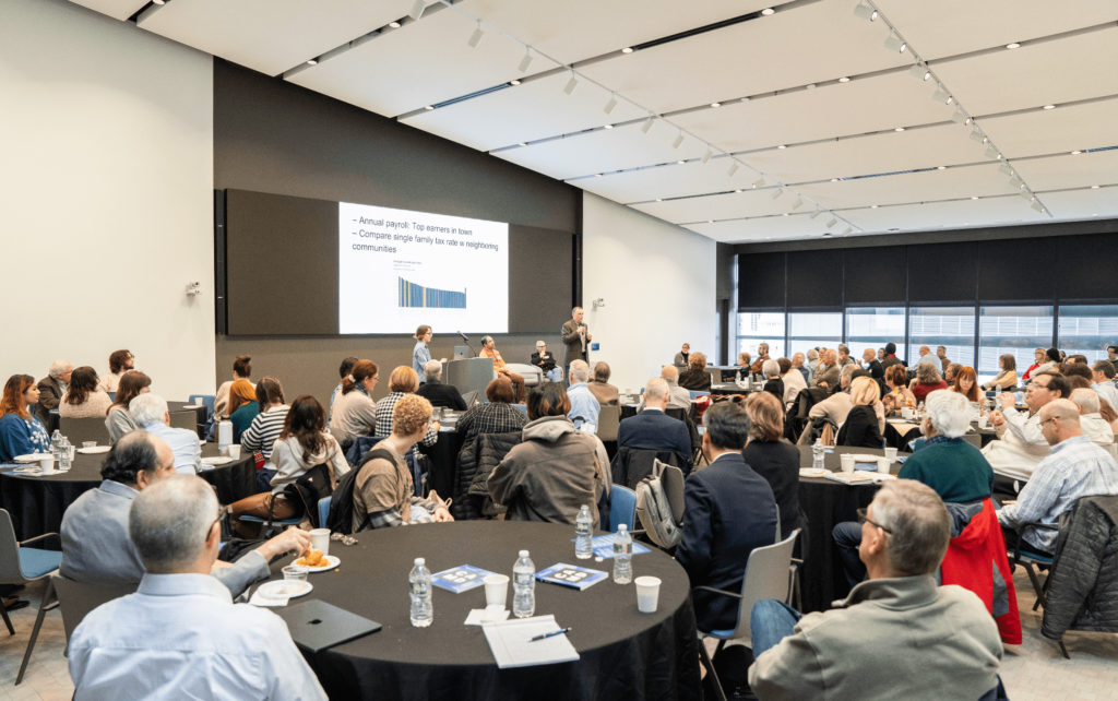 A conference presentation on the future of local news in EXP, Northeastern’s modern event space. A presenter stands at a podium speaking to a seated audience at round tables. The projection screen displays charts and text about 'Annual payroll: Top earners in town' and 'Compare single family tax rate in neighboring communities.' Attendees, mostly middle-aged and older adults, are seated at round tables with water bottles, notepads, and refreshments. The room features track lighting on a white ceiling and dark window shades.