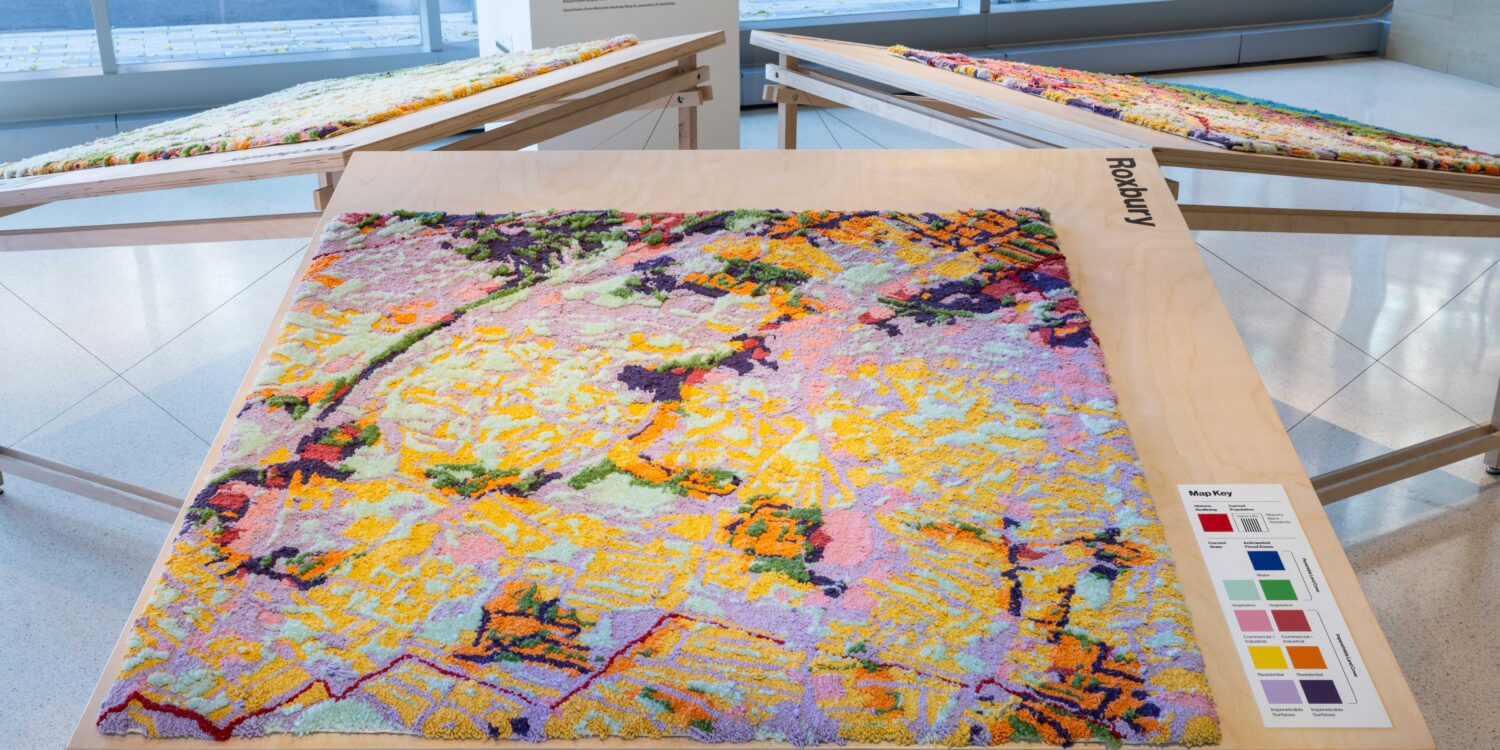Wide angle view of "Soft City" installed in the MIT Museum. "Soft City" is a textile-based art piece depicting a flood zones map of Roxbury in Boston, MA. It is a patchwork of vibrant color including purples, pinks, blue and yellow. The surface is richly textured and soft to the touch. The piece is installed horizontally on a table, giving visitors at the museum an opportunity to touch the art piece.