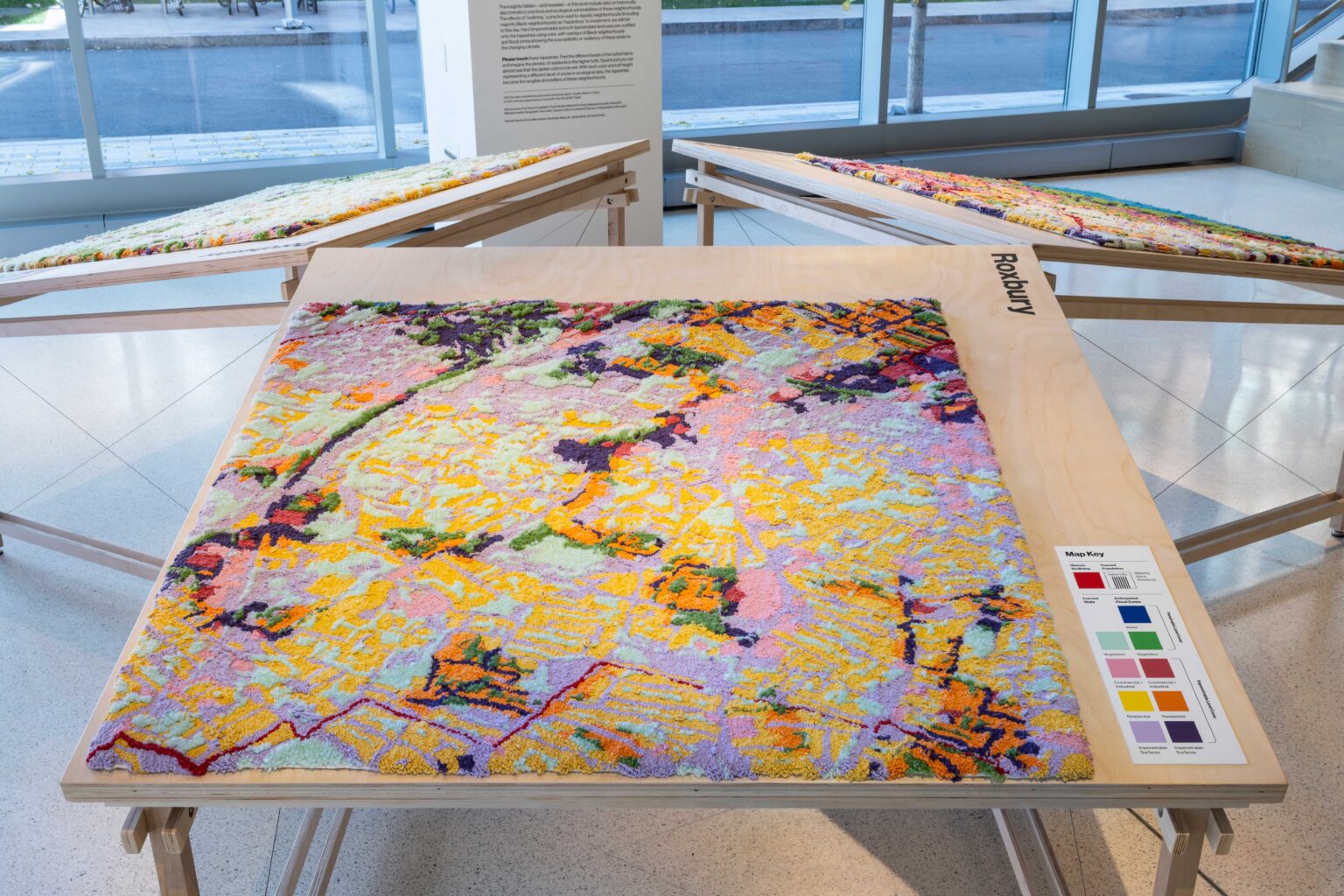 Wide angle view of "Soft City" installed in the MIT Museum. "Soft City" is a textile-based art piece depicting a flood zones map of Roxbury in Boston, MA. It is a patchwork of vibrant color including purples, pinks, blue and yellow. The surface is richly textured and soft to the touch. The piece is installed horizontally on a table, giving visitors at the museum an opportunity to touch the art piece.