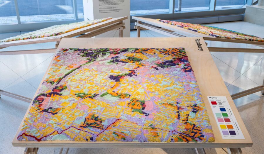 Wide angle view of "Soft City" installed in the MIT Museum. "Soft City" is a textile-based art piece depicting a flood zones map of Roxbury in Boston, MA. It is a patchwork of vibrant color including purples, pinks, blue and yellow. The surface is richly textured and soft to the touch. The piece is installed horizontally on a table, giving visitors at the museum an opportunity to touch the art piece.
