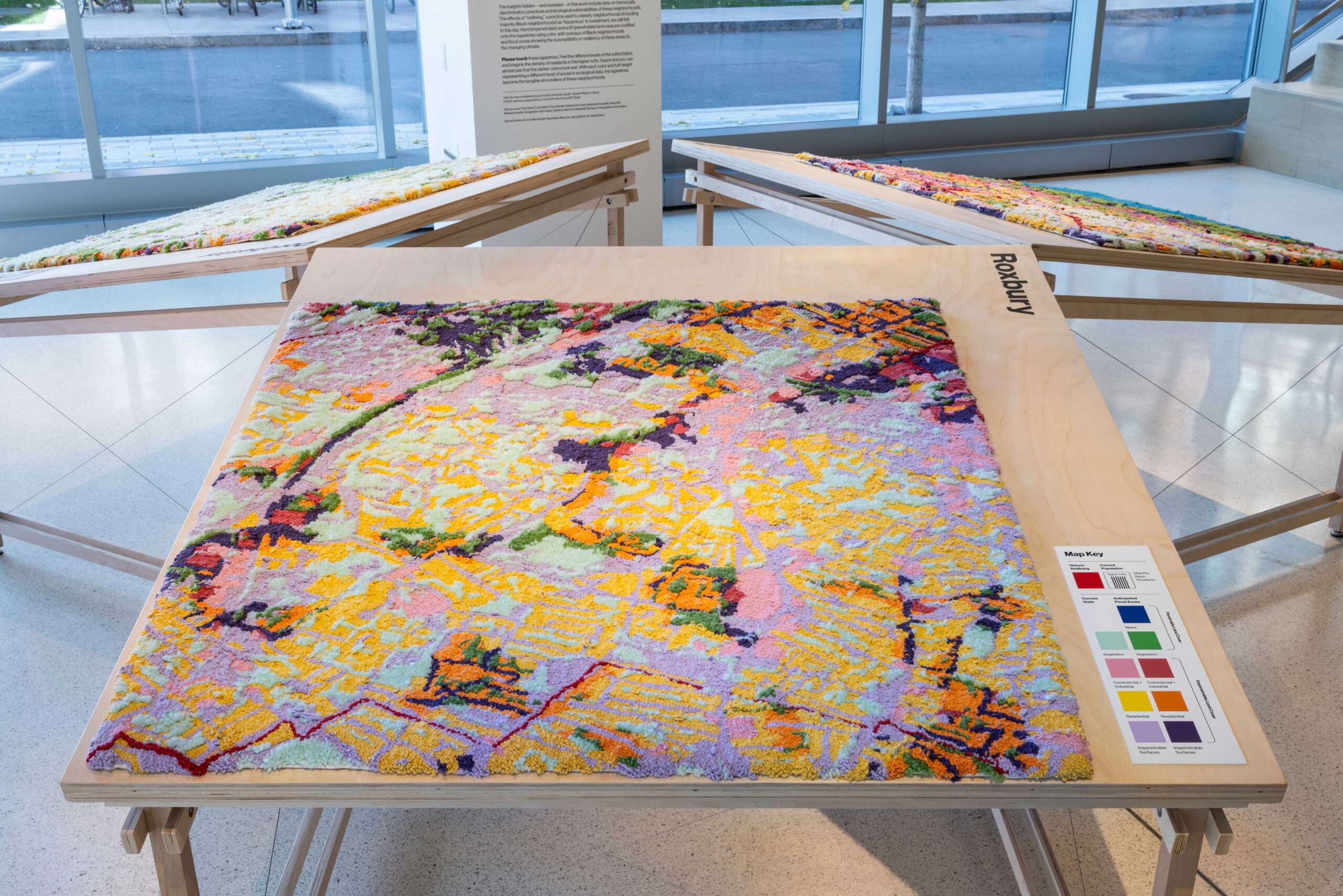 Wide angle view of "Soft City" installed in the MIT Museum. "Soft City" is a textile-based art piece depicting a flood zones map of Roxbury in Boston, MA. It is a patchwork of vibrant color including purples, pinks, blue and yellow. The surface is richly textured and soft to the touch. The piece is installed horizontally on a table, giving visitors at the museum an opportunity to touch the art piece.
