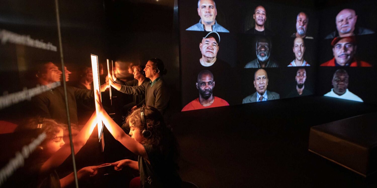 Photo of Rashin Fahandej's artwork, "A Father's Lullaby," installed at the ICA Boston. A big screen of over a dozen peoples' faces is on the right. On the left, exhibition visitors touch glowing screens.