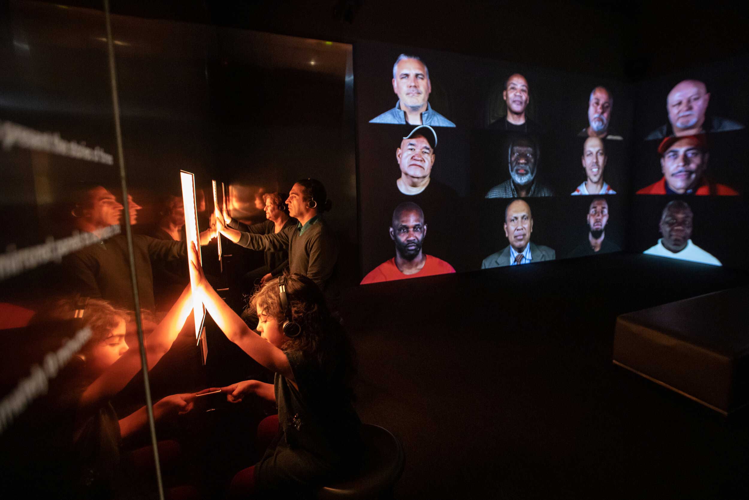 Photo of Rashin Fahandej's artwork, "A Father's Lullaby," installed at the ICA Boston. A big screen of over a dozen peoples' faces is on the right. On the left, exhibition visitors touch glowing screens.