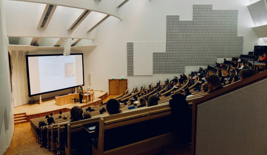 A large university classroom. Huge rows of chairs with a screen and professor at the bottom left of the image.