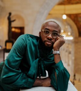 Image of Ph.D. student Caleb Okereke. He has short blonde hair and wears a teal silk shirt with a bow.