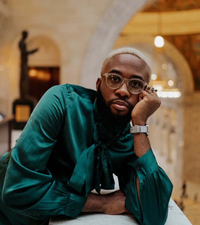 Image of Ph.D. student Caleb Okereke. He has short blonde hair and wears a teal silk shirt with a bow.