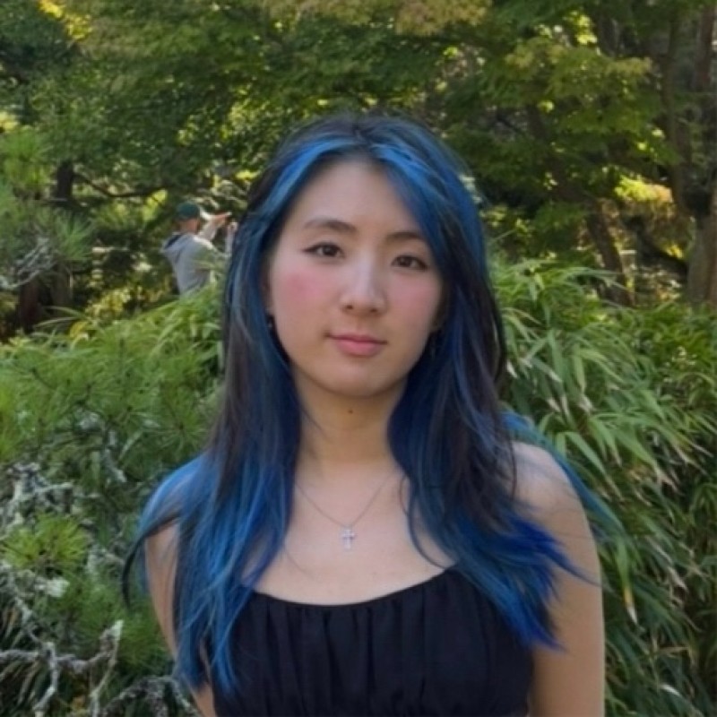 Image of Chelsea, a female student with blue long hair. She is standing in front of greenery/a tree.