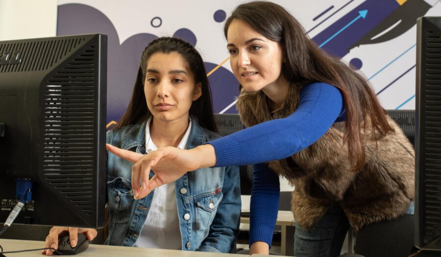 A young woman sitting in front of a computer in an office while another woman standing next to her is pointing at something on her screen. They are both dressed in casual clothes - a bright blue top and furry waistcoat, and a denim jacket.