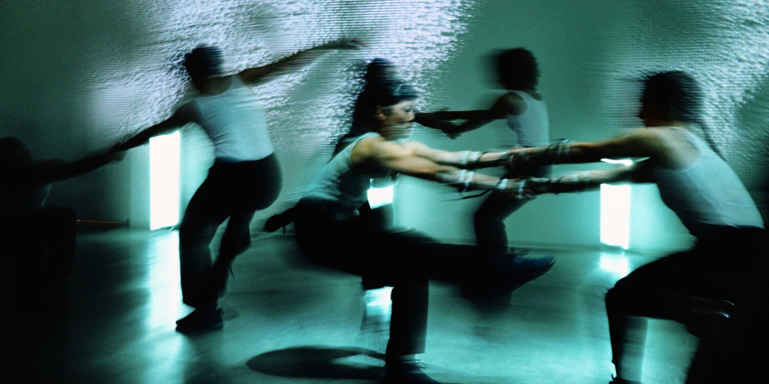 Dreamy, blurred photo of movement artists dancing together. The photo has a green-blue tinge to it; four dancers are fully in the frame and are in pairs of two people holding hands.