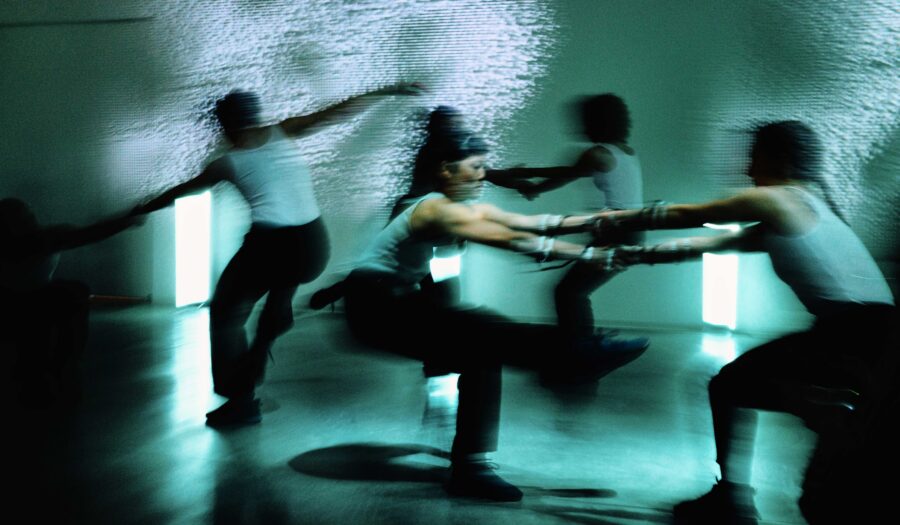 Dreamy, blurred photo of movement artists dancing together. The photo has a green-blue tinge to it; four dancers are fully in the frame and are in pairs of two people holding hands.
