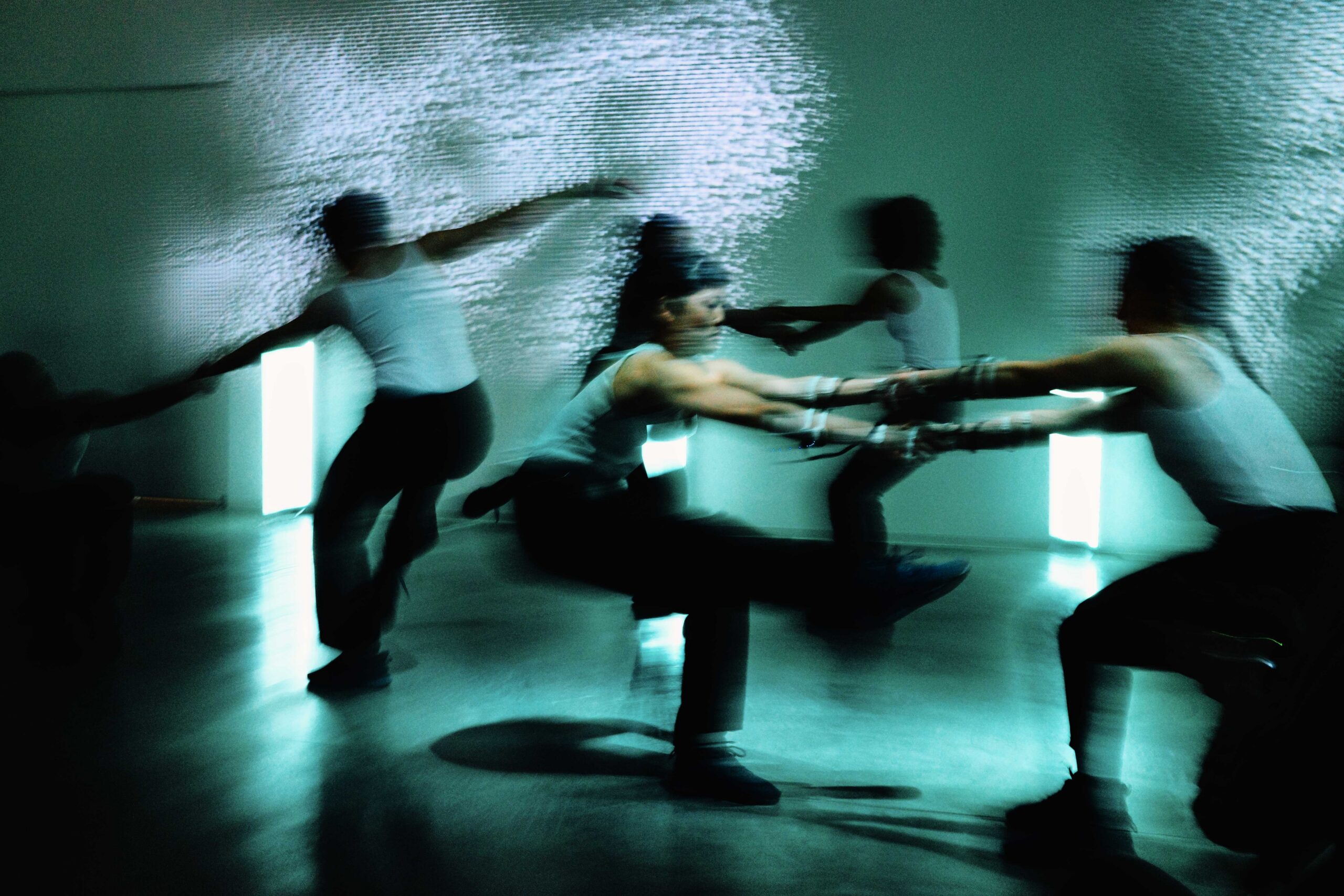 Dreamy, blurred photo of movement artists dancing together. The photo has a green-blue tinge to it; four dancers are fully in the frame and are in pairs of two people holding hands.