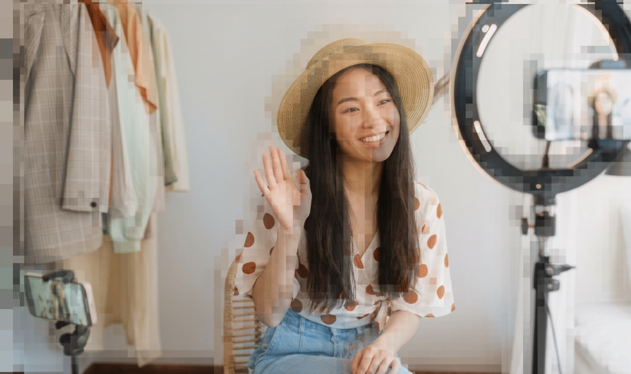 Pixellated image of a female influencer. She is an Asian woman smiling and waving hello in front of a ring light.