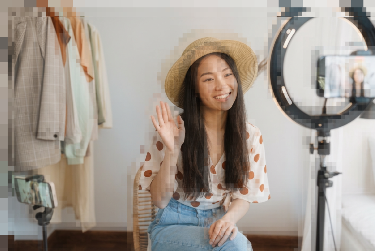 Pixellated image of a female influencer. She is an Asian woman smiling and waving hello in front of a ring light.