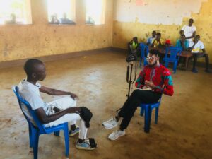 Caleb Okereke sits across from a man in a town that's part of Uganda’s Bidi Bidi refugee settlement. They are sitting in blue plastic chairs in a mostly unfurnished room. A table with six onlookers is behind them; they are watching the interview. 